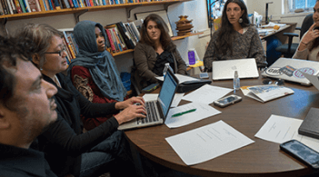students and professor seated at a round table with computers, books and paper