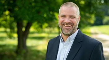 Photo of male faculty member smiling and standing outside in front of a tree.