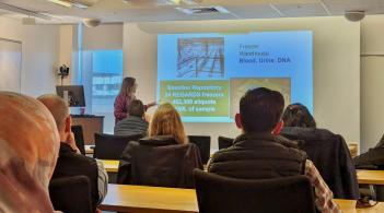 A researcher is standing at the front of a conference room, giving a presentation to a seated audience.