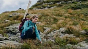 A student crouching on a mountainside looking at plant growth, she is wearing a backpack with equipment in it.