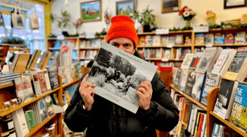 A man in an orange beanie hat stands in a bookstore holding up the book "Vermont Female Farmers" over his face.