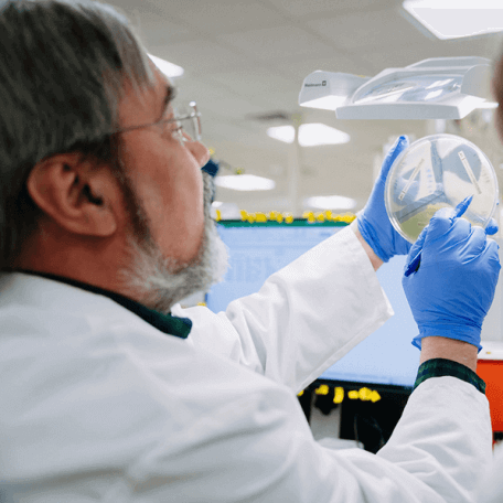 A man in a lab coat holds a petri dish up to the light