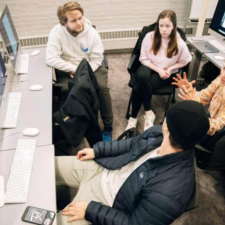 A group of students sitting around a desk of computers having a discussion