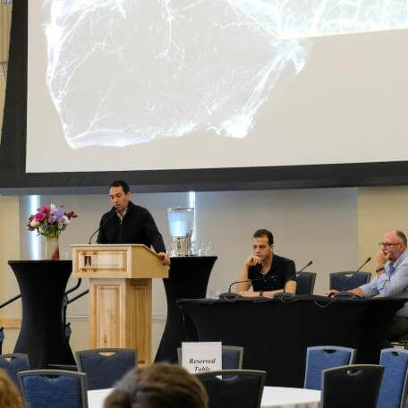 A researcher delivers a presentation at a podium in front of a large display screen, while two additional researchers are seated at a table to the side linking to Events Page