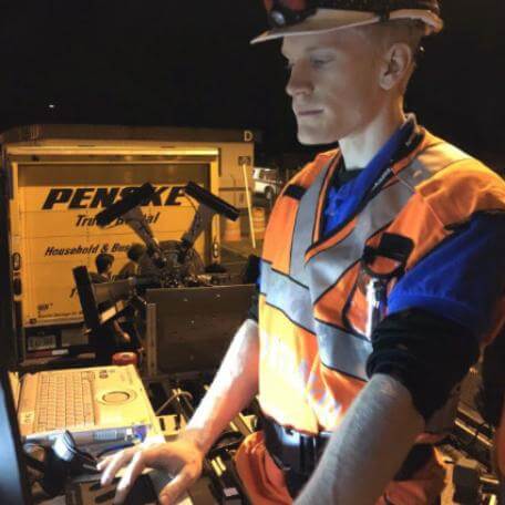 A man wearing an orange safety vest and glasses is focused on working on a laptop outdoors
