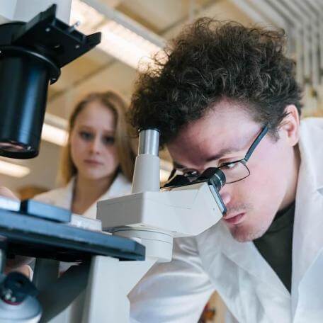 White-coated scientist looks through mictoscope with colleague behind