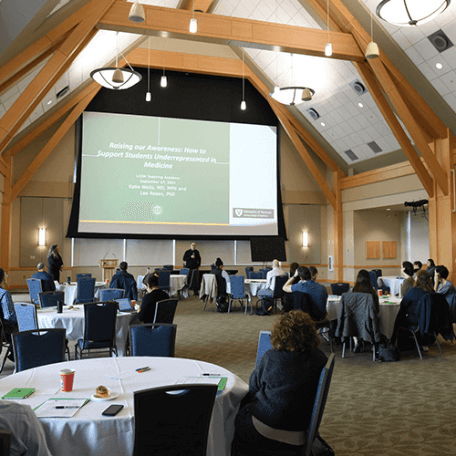 A group of people sitting at tables in a large room with a presentation on screen