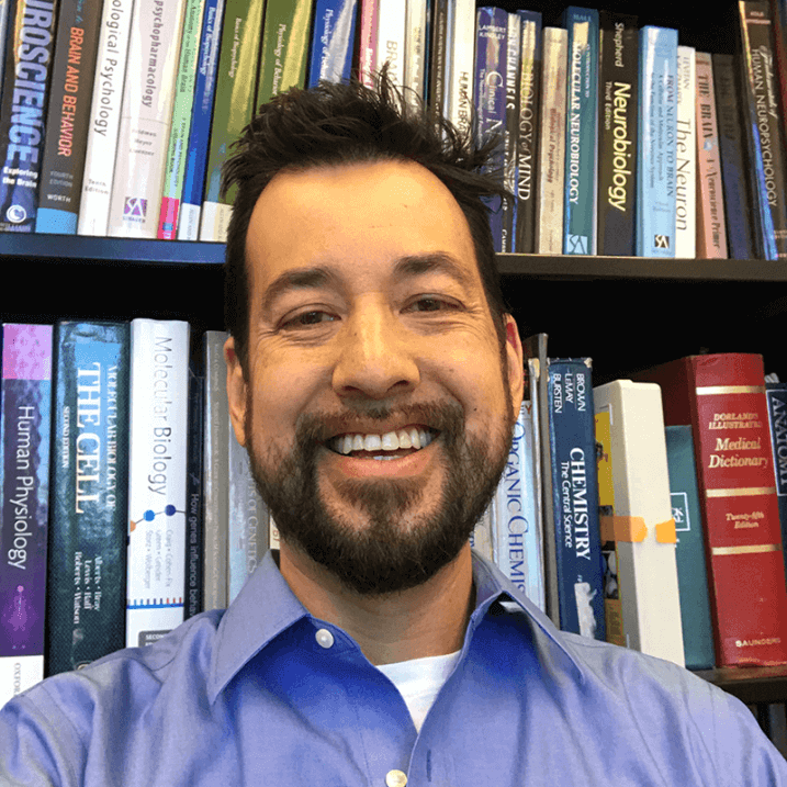 Jom, with a full face smile, in an office setting with book lined shelves in the background
