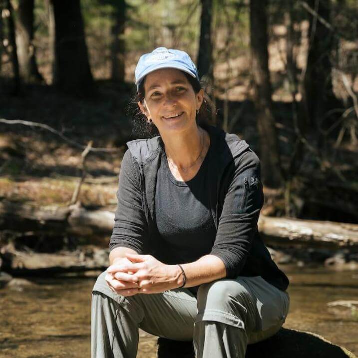 Carol Adair outside working near a stream, smiling to camera