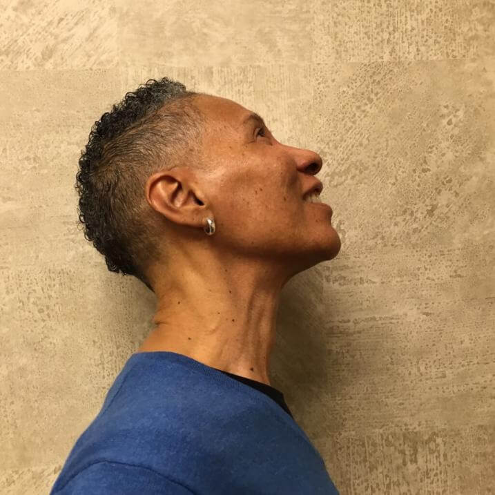 profile view of sharon, wearing blue shirt standing in front of textured beige wall looking upwards toward the light!