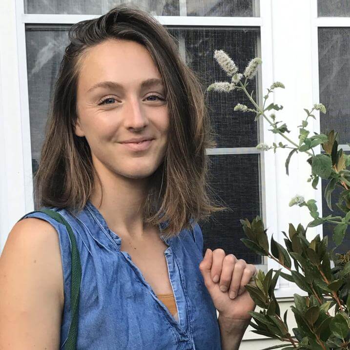 Margaret, wearing a blue dress and with her brown hair loose, smiling and standing next to a head-high mint plant in front of an elder's home