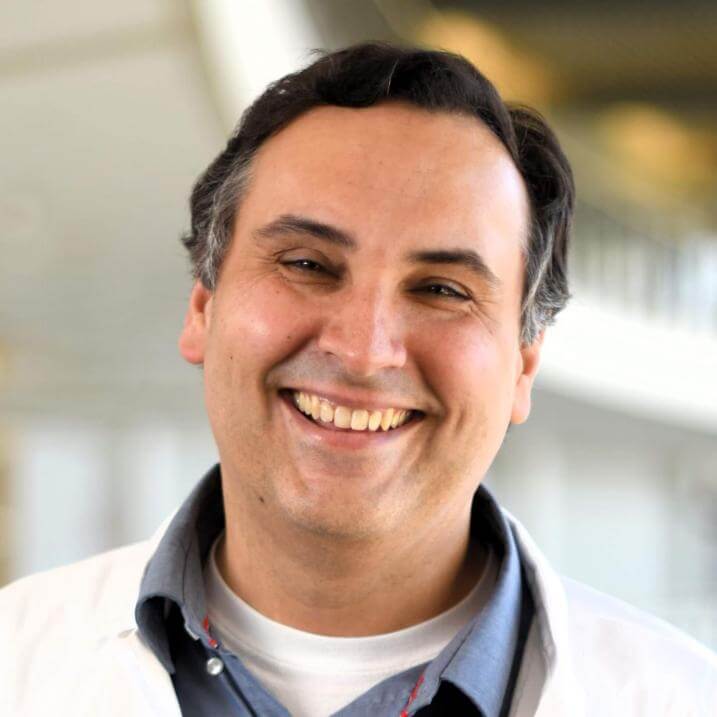 Headshot of a researcher and doctor in a white coat against a blurred indoor background.