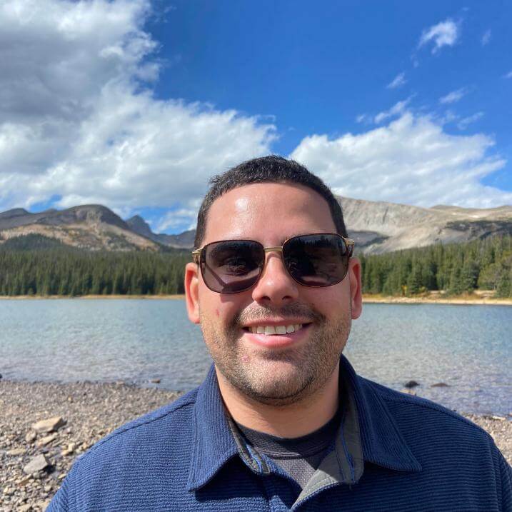 Headshot of Dale Henderson. Dale is wearing a blue shirt and sunglasses. There is water and blue sky behind him.