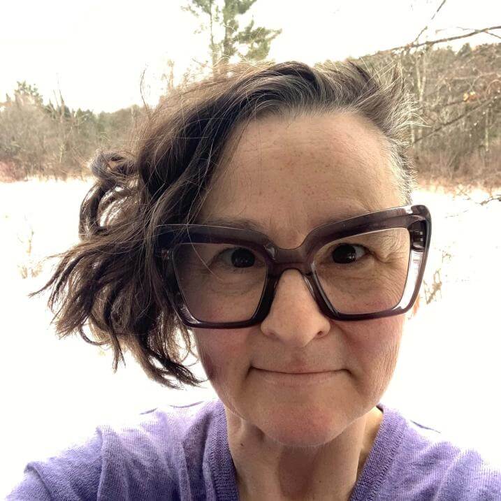 A non-binary white person in their forties, with dark hair worn loose and wild to one side, and large, heavy-framed glasses