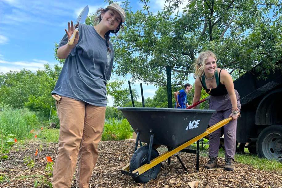 Two PBIO 290 students holding a trowel and pushing a wheel barrow in the Tommy Thompson Community Garden pollinator habitat area.