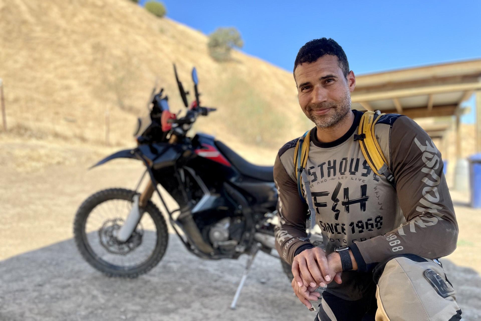 man with dark hair kneeling in front of black and red motorcycle