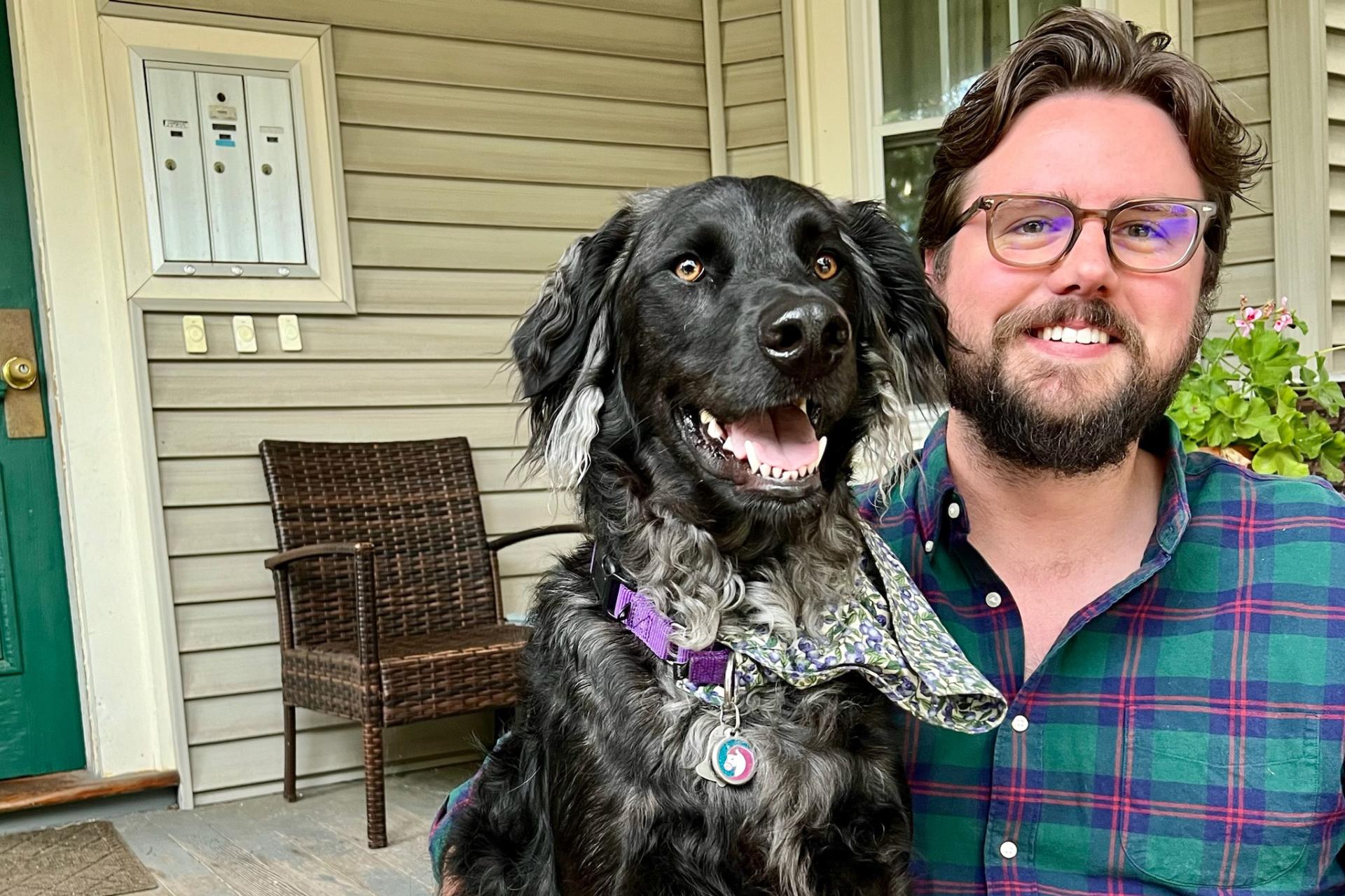 man with glasses and dark hair and beard, wearing plaid shirt, next to black dog on porch of house