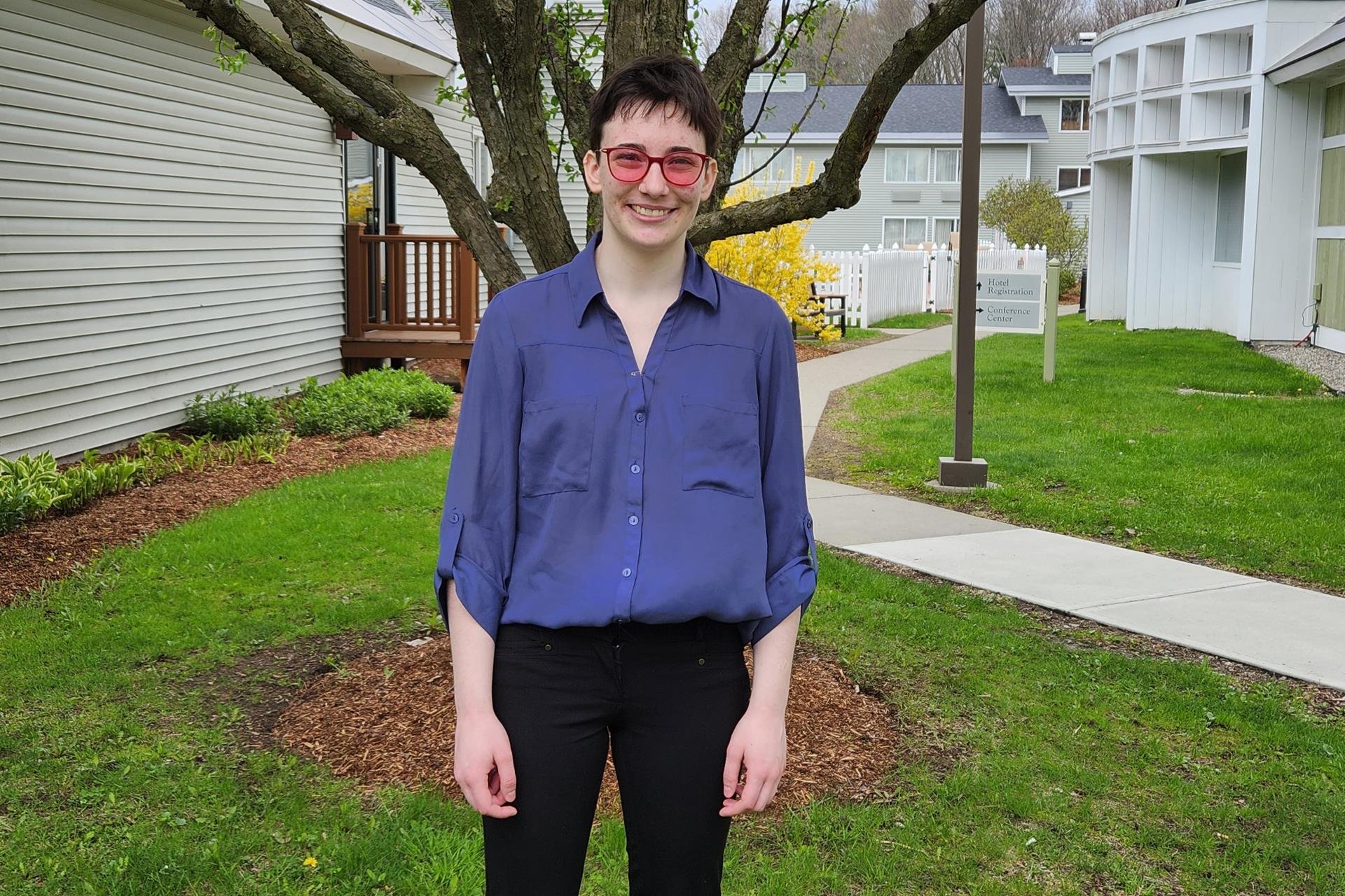 young woman with short hair, blue shirt, and rose glasses standing in front of tree