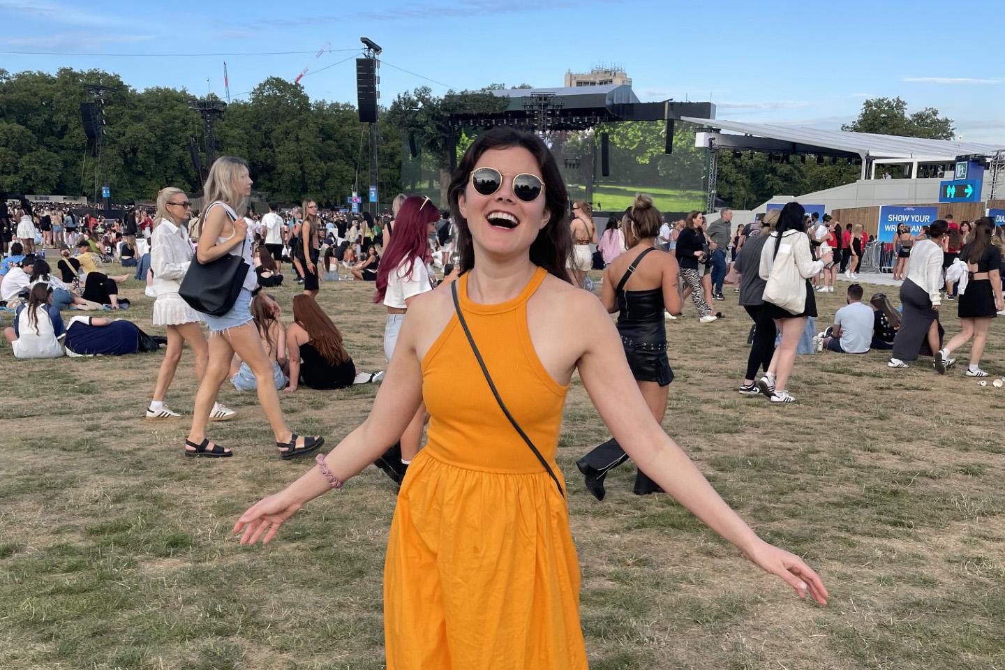woman with long brown hair and sunglasses in yellow dress smiling in middle of field surrounded by people