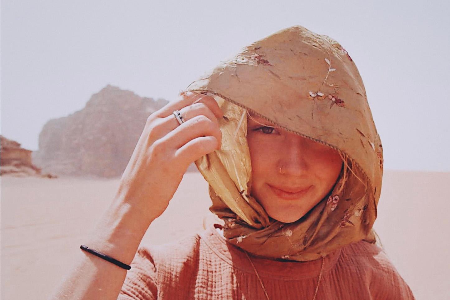 young woman in beige headscarf standing in desert with rock formations in background