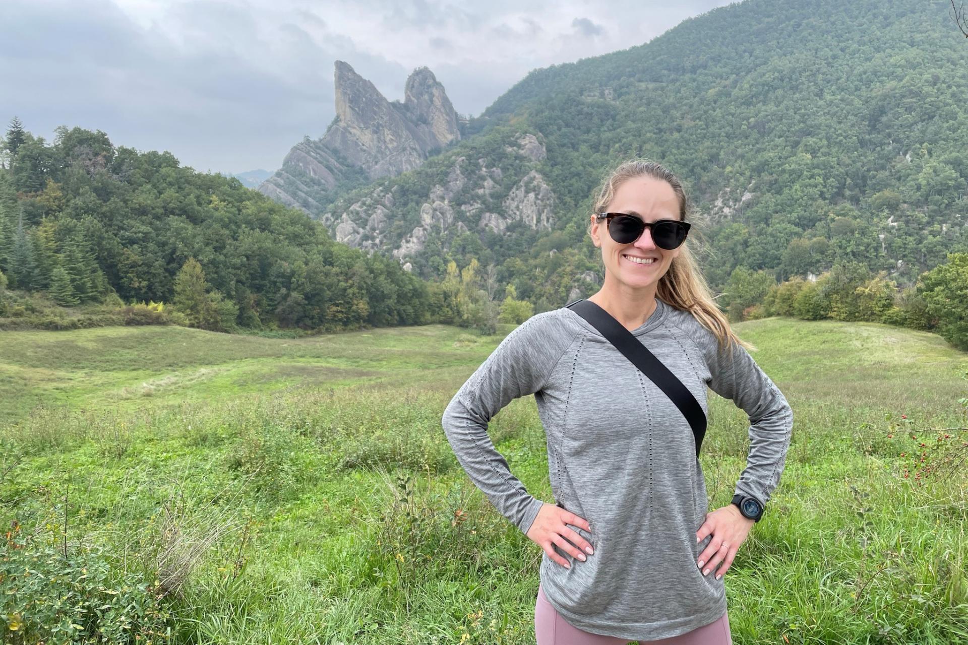 woman with blond ponytail and sunglasses standing in green field with mountains in background