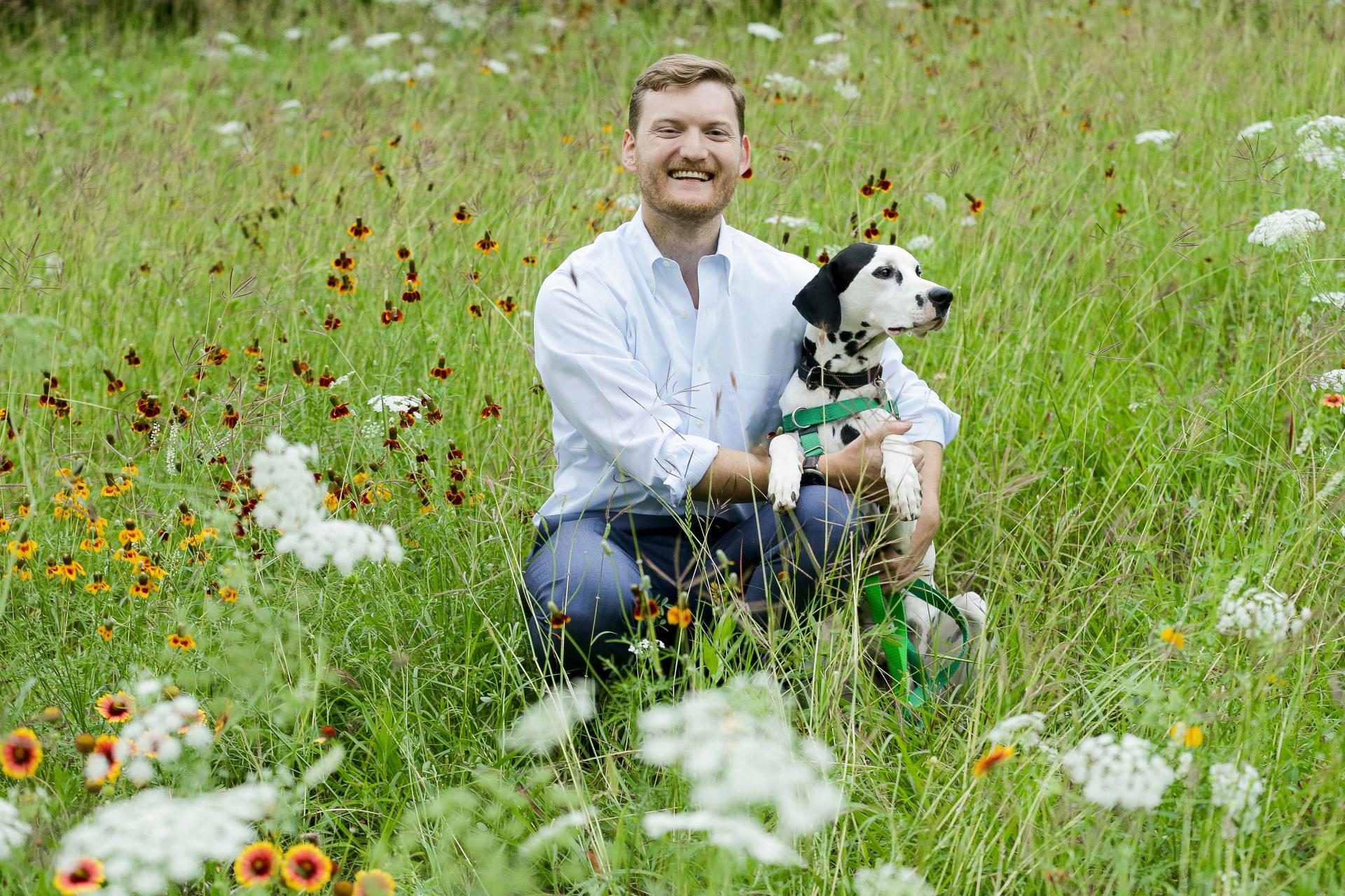man with beard in white shirt and jeans crouching in grassy field with wildflowers next to black and white dog