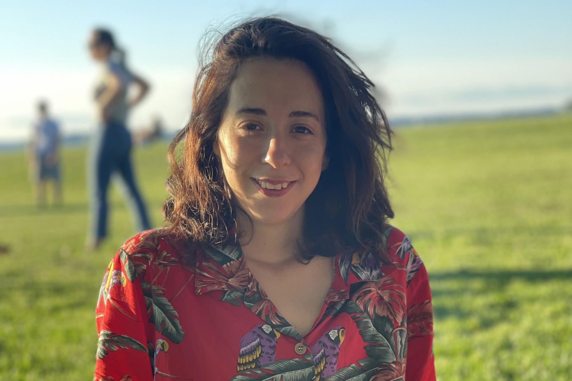 young woman with long brown hair and red shirt sitting in grassy field with other people in background