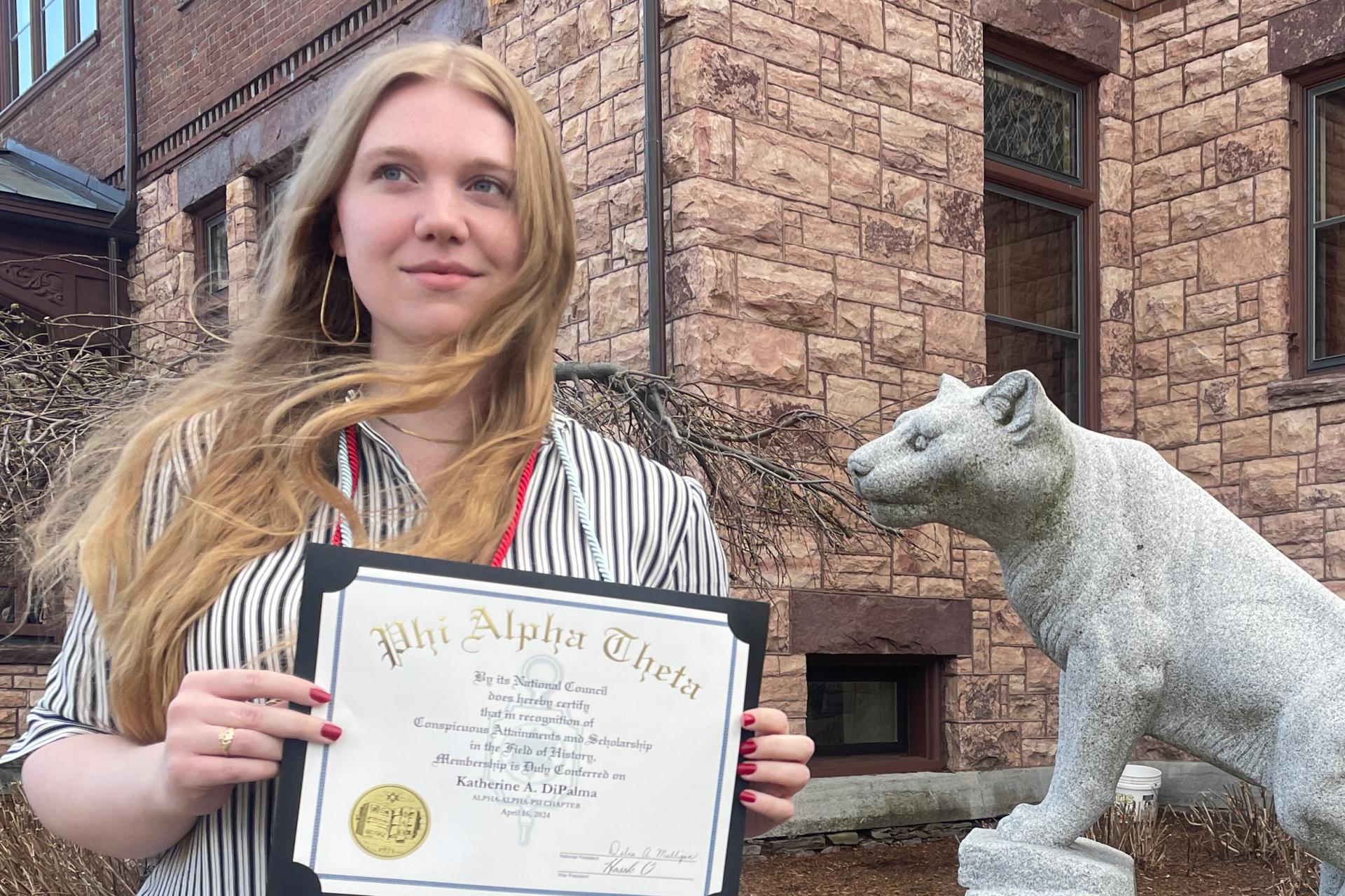 young woman with long blonde hair wearing striped shirt and standing in front of stone building holding Phi Alpha Theta membership certificate