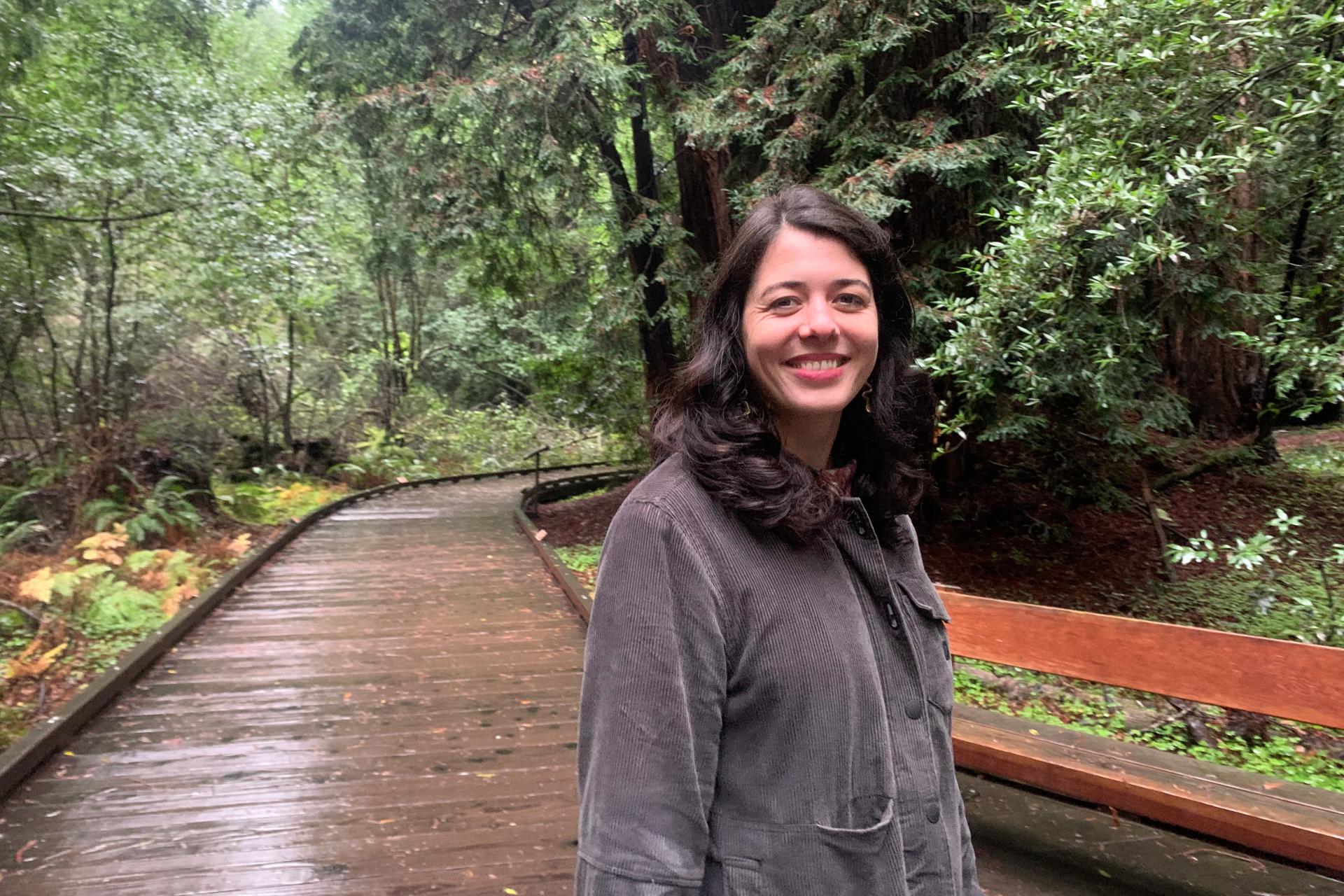 woman with long brown hair and gray jacket standing on wooden boardwalk in the woods