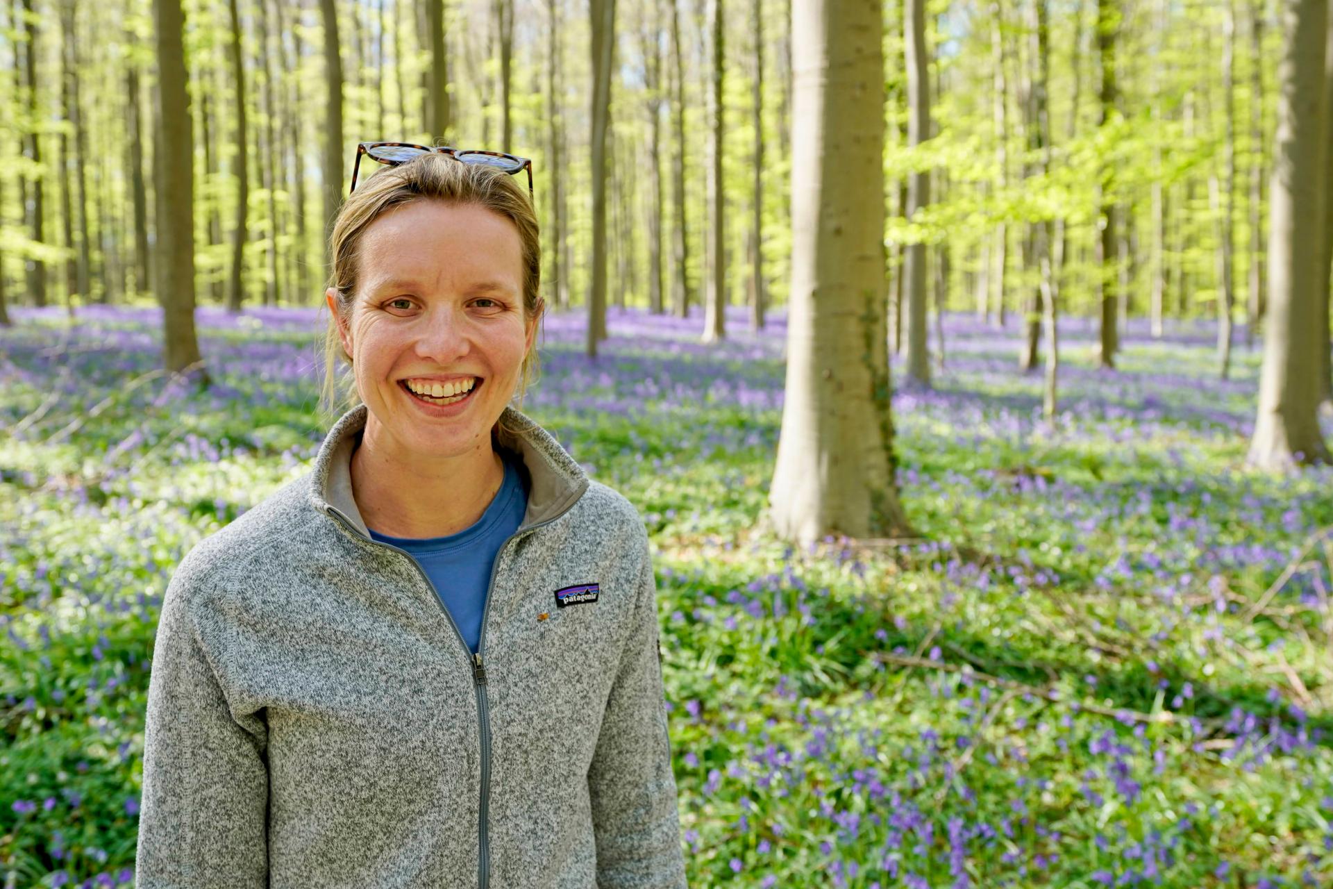 woman with blond hair and gray zip-up sweater standing in sunny forest with purple flowers