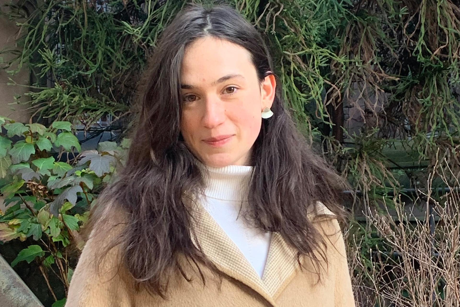 young woman with long dark hair in white turtleneck and tan coat standing in front of wall with greenery