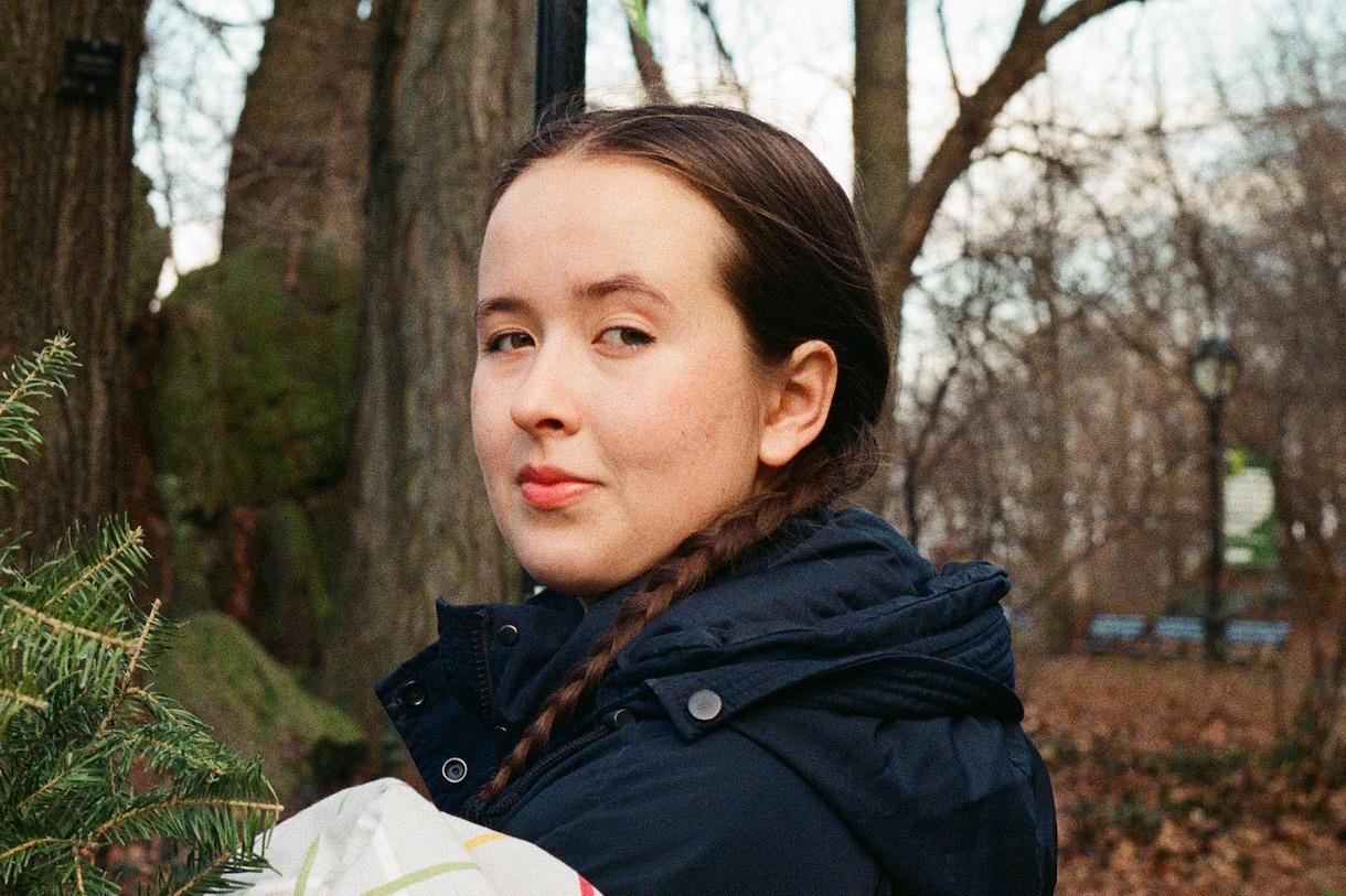 Student Nora Sissenich in blue jacket standing in woods by a tree