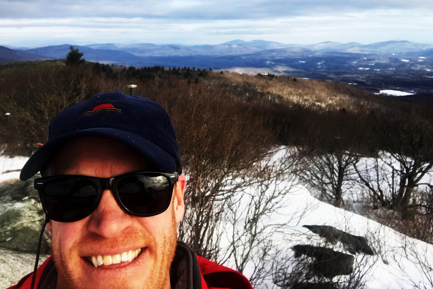 close-up of man in baseball cap and sunglasses with partly snow covered mountains behind