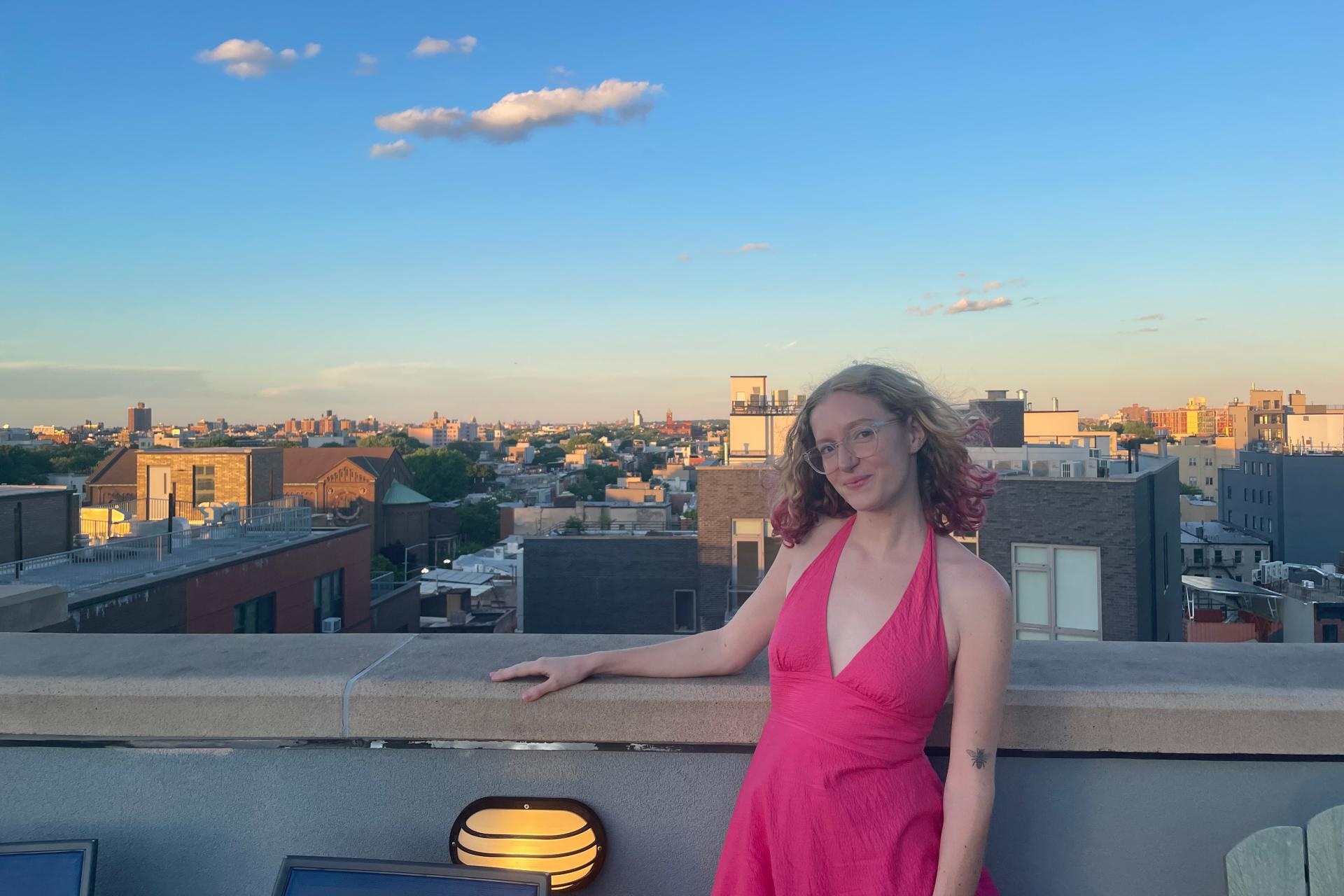 young woman with long, light hair and pink dress standing by wall overlooking city