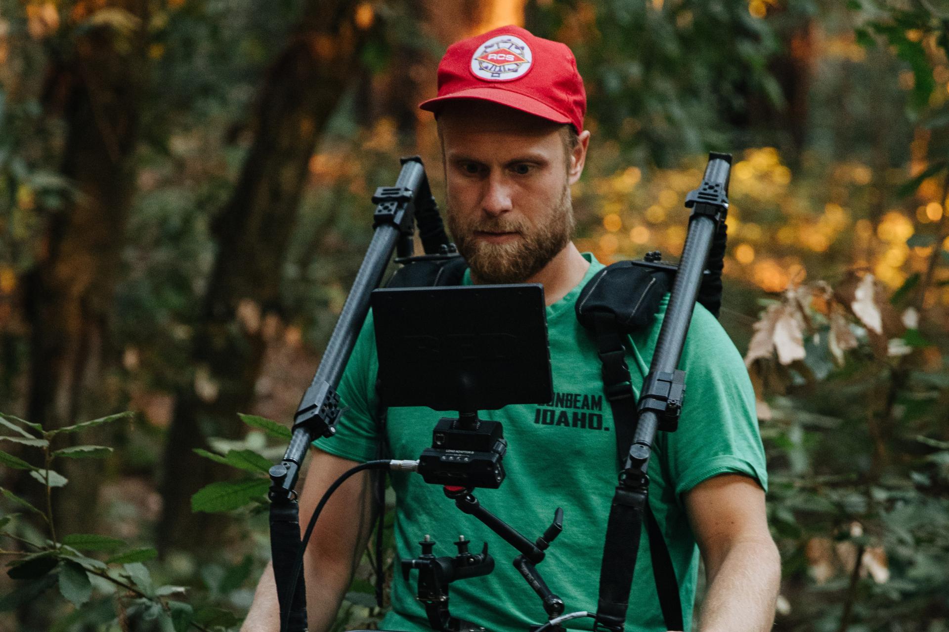 bearded man wearing green t-shirt and red hat shoots a film in the woods