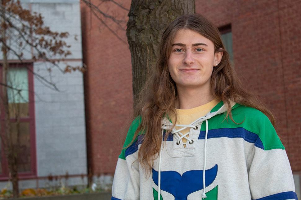 young person with long light brown hair and light gray/blue/green sweatshirt standing by tree outside classroom building