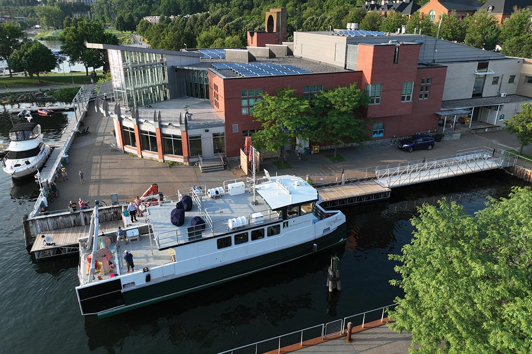 a white and green vessel sits docked in the water in front of a large brick building