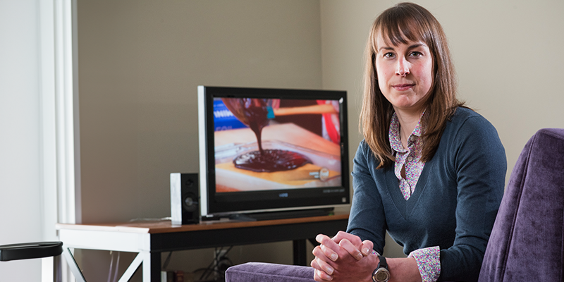Lizzy pope sitting in a chair turned towards the camera with her hands clasped and a tv in the background