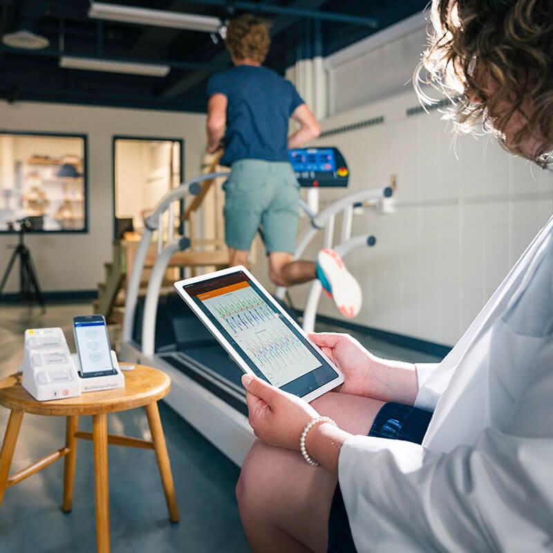 a person holds a ipad with data on it as another person with sensors on them runs on a treadmill