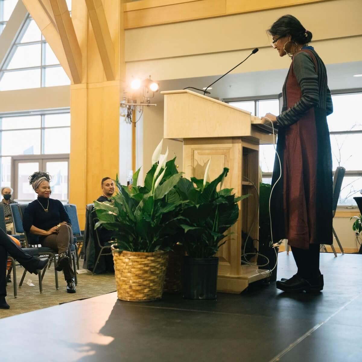 a woman standing at a lectern speaking to a group of seated people