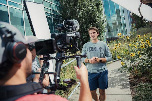 Behind the shoulder of a camera operator filming a student speaking