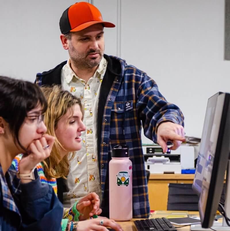 Students in front of a computer are directed by an instructor.