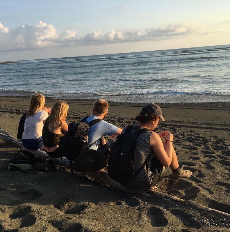 Five students sitting on a piece of wood on a beach