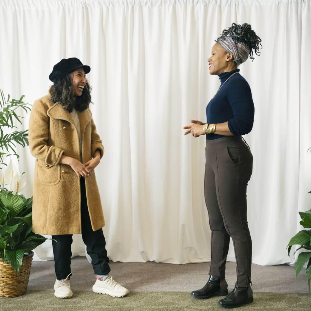 Kristy Drutman and Majora Carter looking at each other laughing in front of a white background with plants on either side of them.