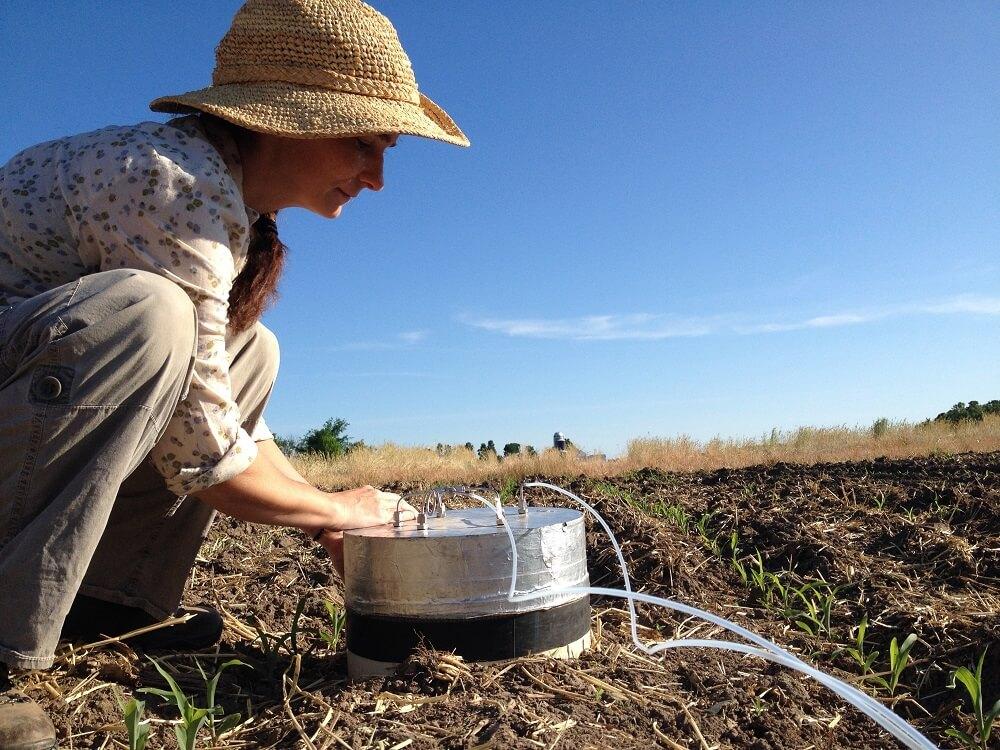 A person in a straw sunhat is squatting beside a metal piece of scientific equipment in a field. There is blue sky in the background.
