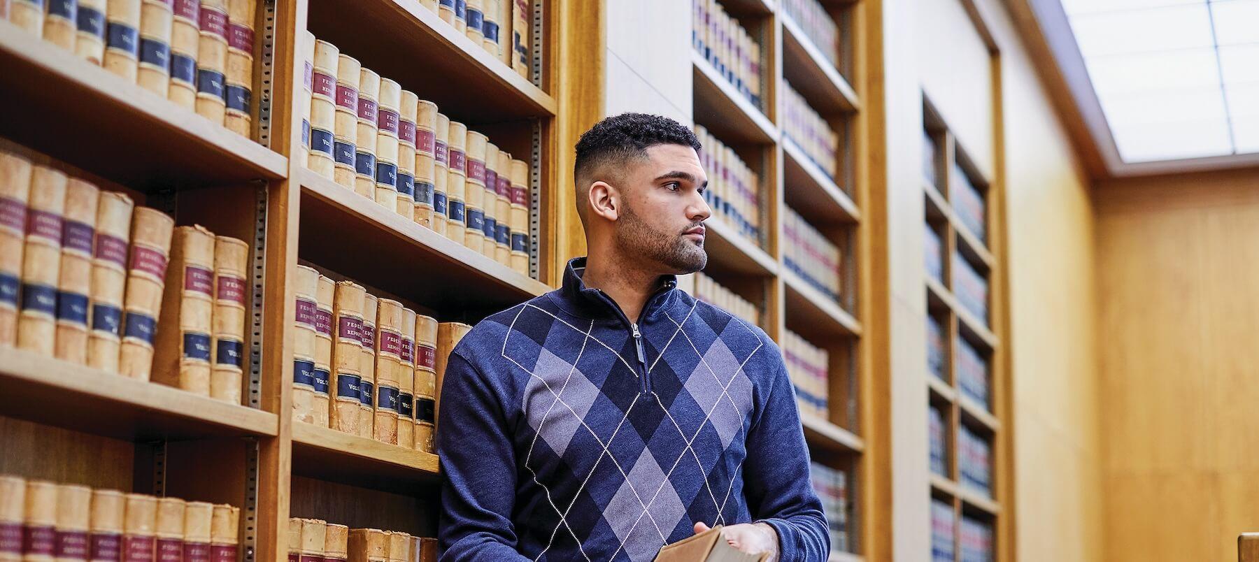 A young man in an argyle sweater stands next to a large shelf filled with books.