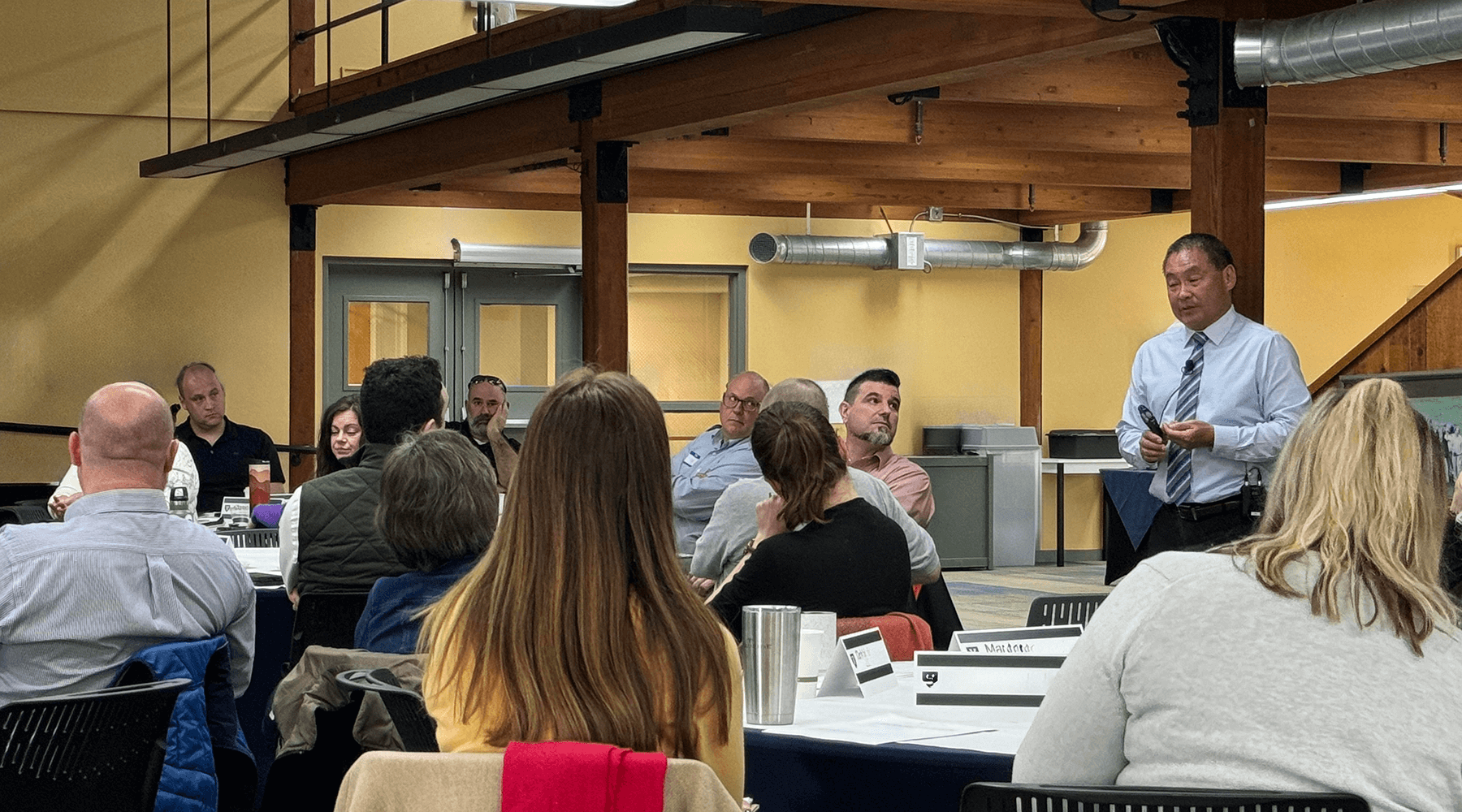 Presenter standing while talking to a room a people sitting at round tables during a training
