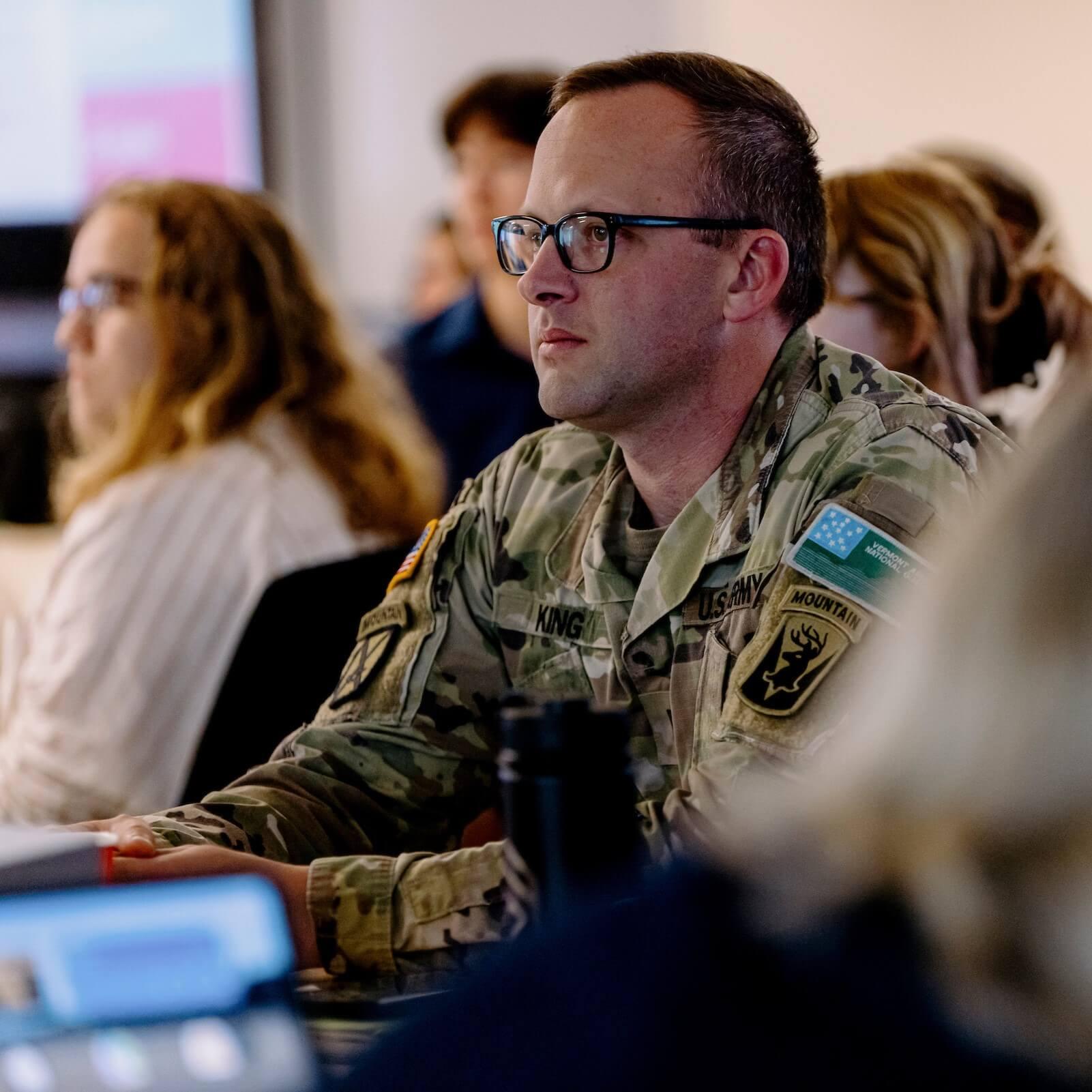 Man in Vermont National Guard uniform sitting at a table, listening attentively to someone off camera