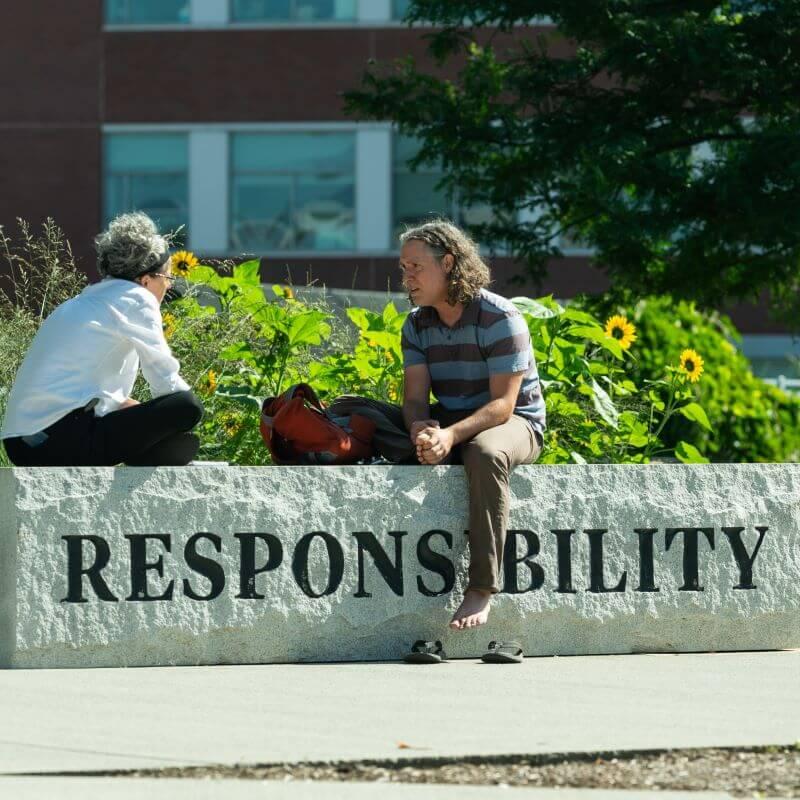 Employees outside the Davis Center sitting on one of the Stone benches labeled "Responsibility" while conversing.