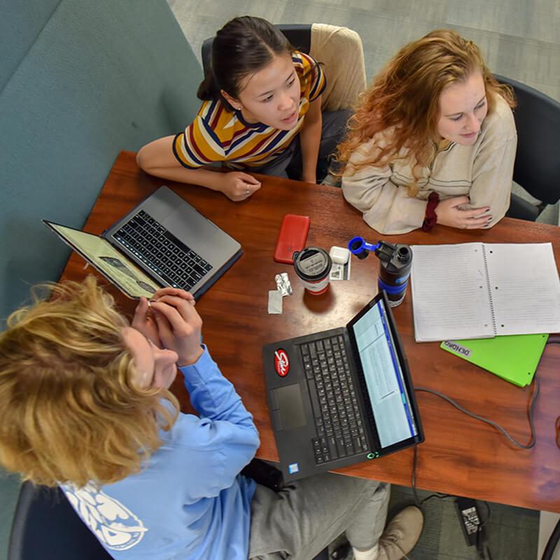 Students sitting at a table looking in the same direction with their laptops open.